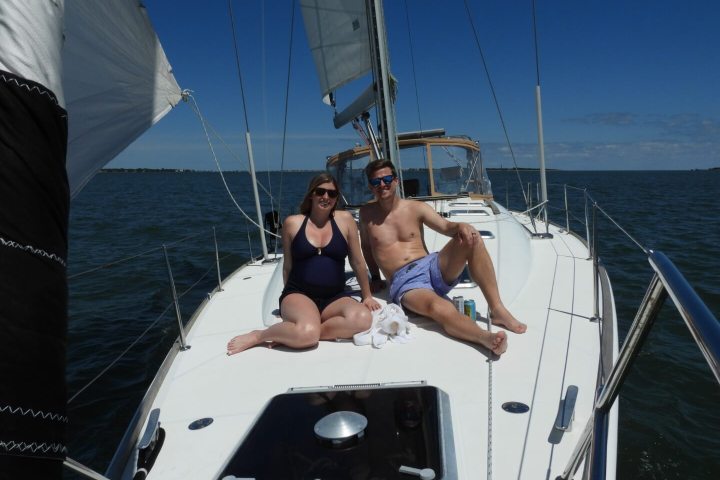 Two people relax on a sailboat deck in swimsuits under a clear blue sky.