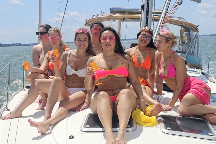 Group of six women in swimsuits sitting on a sailboat deck, holding drinks.