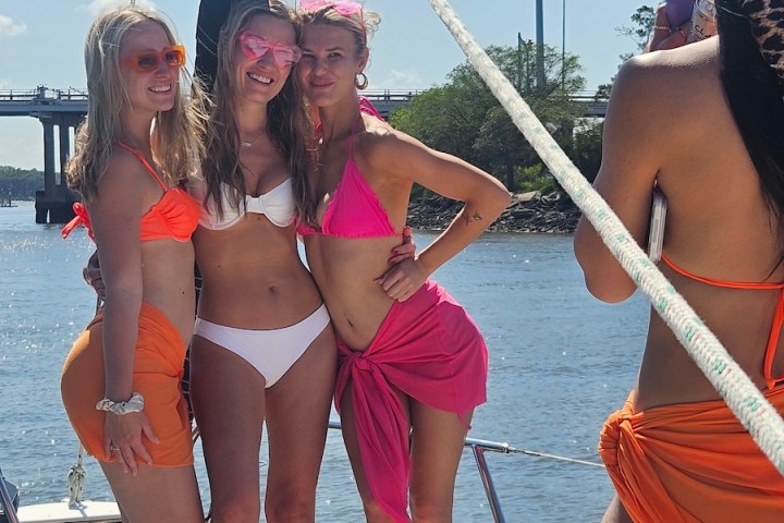 Three women in swimsuits pose on a boat with a bridge in the background.