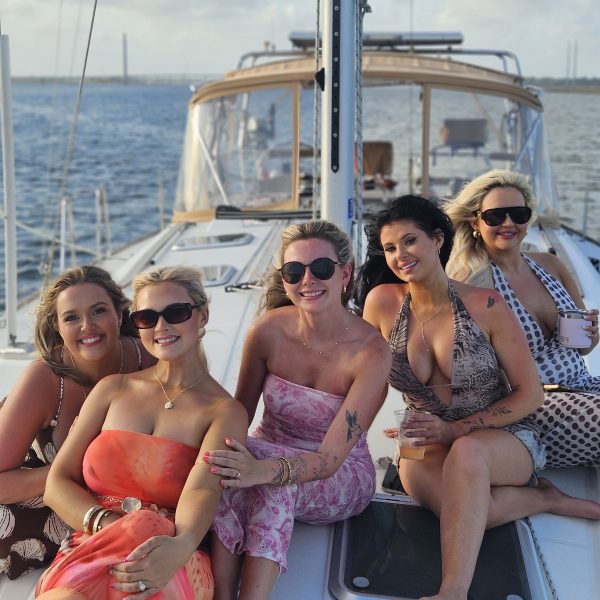 Five women in summer dresses and sunglasses sitting on a sailboat.