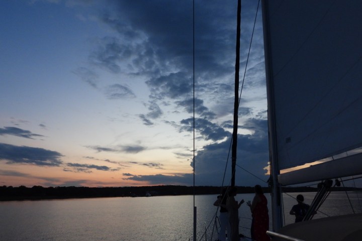 Sailboat on calm water at sunset with people on deck and a cloudy sky.