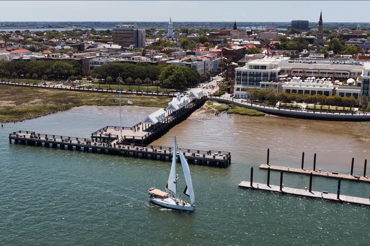 A sailboat near a city shoreline with piers and buildings visible.