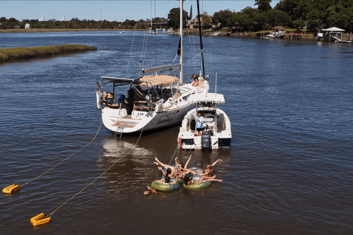 Sailboat on river with people relaxing on float tubes nearby.