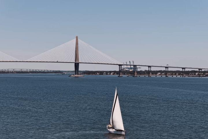 Sailboat on water with a suspension bridge in the background under clear skies.