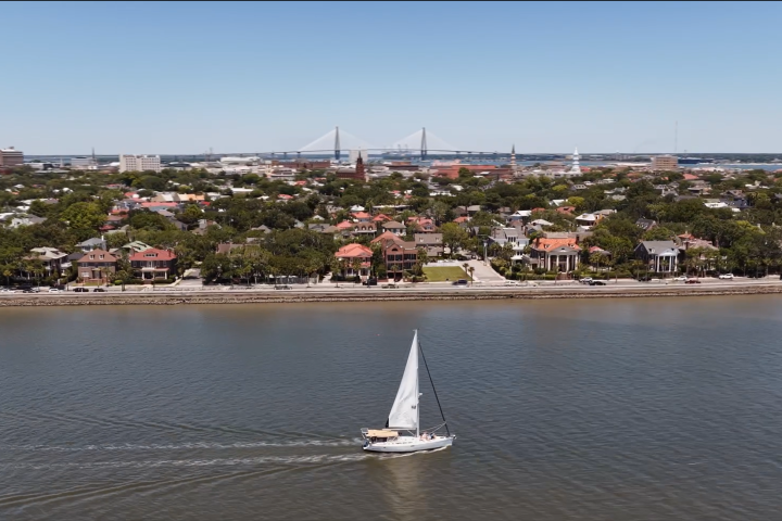 Sailboat on river with cityscape and bridge in background under a clear sky.