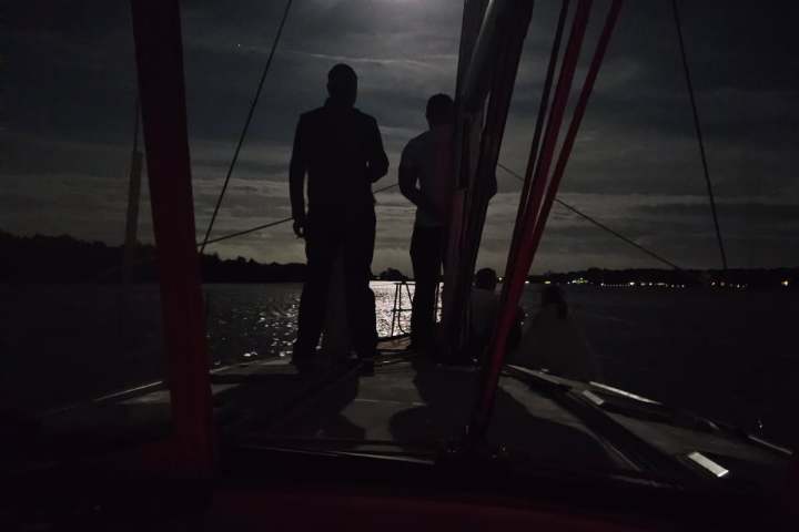 Silhouetted figures on a sailboat under a bright moonlit night sky.
