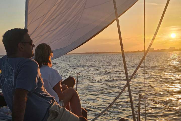 Two people relax on a sailboat at sunset, with the ocean and sun in the background.