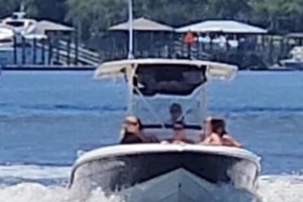 Speedboat with people cruising on a river, trees and docks in background.