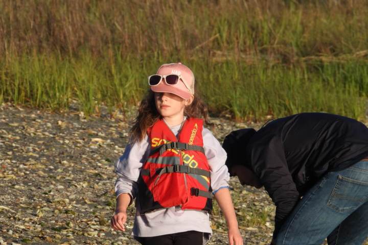 Child in life vest and hat walks on rocky shore.