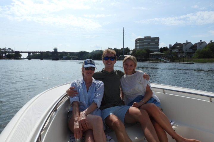 Three people sitting on a boat near a shoreline on a sunny day.