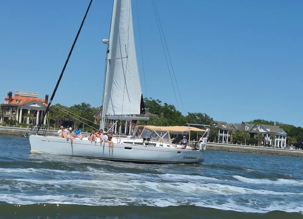Sailboat with people on deck, sailing near shoreline homes on a sunny day.