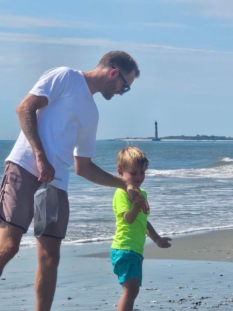 Man and child walking on a beach with a lighthouse in the distance.