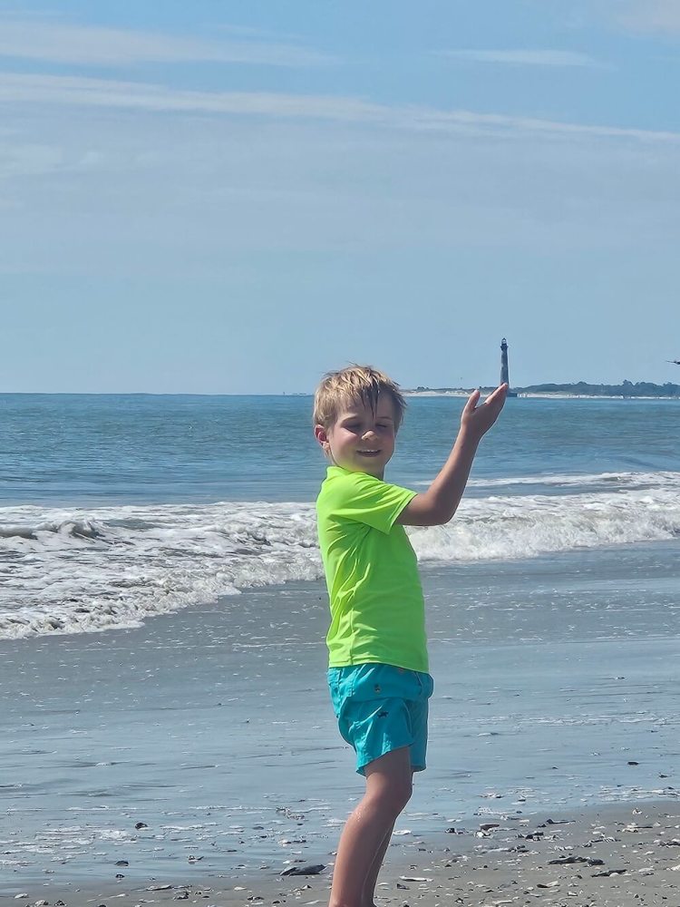 Child on beach holding up hand towards distant lighthouse.