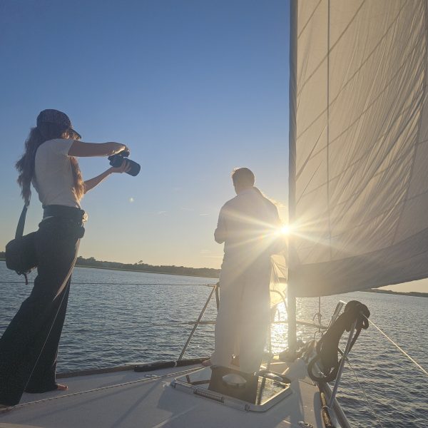 Two people on a sailboat at sunset, one photographing while sun shines through sail.